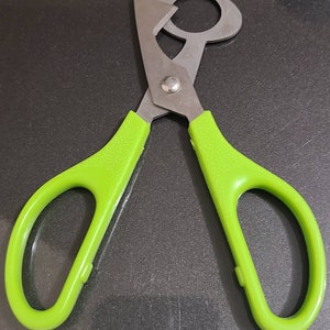 May include: A pair of scissors with a unique design, featuring a silver blade and bright green handles. The blades have a circular opening and a pointed tip. The handles are textured for grip.