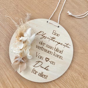 Könnte beinhalten: Ein rundes Holzschild mit einer cremefarbenen Schnur zum Aufhängen. Das Schild hat deutschen Text und ist mit Trockenblumen und einer Schleife verziert. Der Text lautet: "Eine Physiotherapeutin der man blind vertrauen kann. Von -en Danke für alles!"