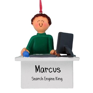 Puede incluir: Un adorno navideño personalizado que representa a un hombre de dibujos animados sentado en un escritorio con una computadora, un ratón y un teclado. El adorno es blanco con el nombre "Marcus" y el texto "Search Engine King" impreso en él.
