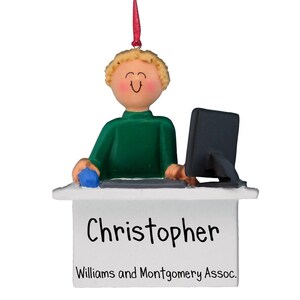 Puede incluir: Un adorno navideño personalizado que representa a un hombre de dibujos animados con cabello rubio sentado en un escritorio con una computadora. El adorno es blanco con el nombre "Christopher" y el texto "Williams and Montgomery Assoc." impreso en él.