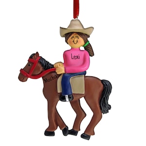 May include: A personalized Christmas ornament featuring a girl wearing a brown cowboy hat and pink shirt riding a brown horse named Mocha. The ornament is decorated with the girl's name, Lexi.