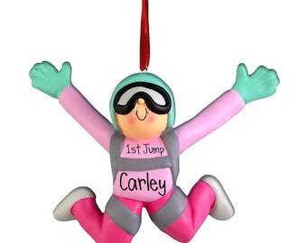 Può includere: Un ornamento da appendere a forma di paracadutista rosa e grigio con un casco e occhiali verdi. L'ornamento ha il testo "1st Jump" e "Carley".