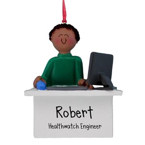Puede incluir: Un adorno personalizado que representa a un hombre de dibujos animados con piel morena sentado en un escritorio con una computadora. El adorno es blanco con el nombre "Robert" y el título "Healthwatch Engineer" escrito en él.
