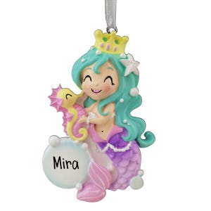 Puede incluir: Adorno de sirena caprichoso con corona y caballito de mar. La sirena tiene cabello turquesa, cola morada y rosa, y una estrella de mar blanca. El nombre "Mira" está en una burbuja blanca. Cuelga de una cinta plateada.
