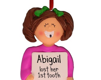 Può includere: Un ornamento natalizio personalizzato con una ragazza dai capelli castani e una camicia rosa che tiene un cartello che dice "Abigail ha perso il suo primo dente".