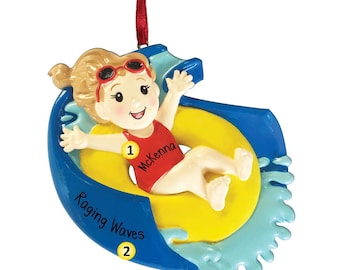 Op de afbeelding: Een gepersonaliseerde kerstversiering met een meisje in een rode badpak dat op een gele binnenband een blauwe waterglijbaan afdaalt. De versiering heeft de tekst "Raging Waves" en "Mckenna" erop.