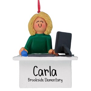 Puede incluir: Un adorno navideño personalizado que representa a una mujer de dibujos animados con cabello rubio sentada en un escritorio con una computadora. El adorno es blanco con el nombre "Carla" y el texto "Brookside Elementary" escrito en él.