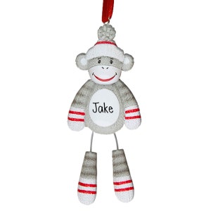 May include: A gray sock monkey ornament with red and white striped accents. The monkey wears a white hat with a red stripe and a gray pom-pom. The name "Jake" is printed on the monkey's chest. A red ribbon is attached for hanging.