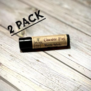 May include: A black lip balm tube with a label that reads "Chocolate Frog Kettle Works." The label has a vintage design with a bee illustration. The text "2 PACK" is printed in black diagonally across the top of the image.