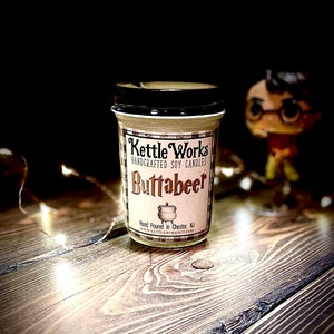 May include: A glass jar candle with a black lid and a white label that says "Kettle Works Handcrafted Soy Candles Buttabeer" and "Hand Poured in Chester, NJ www.kettleworksco.com".
