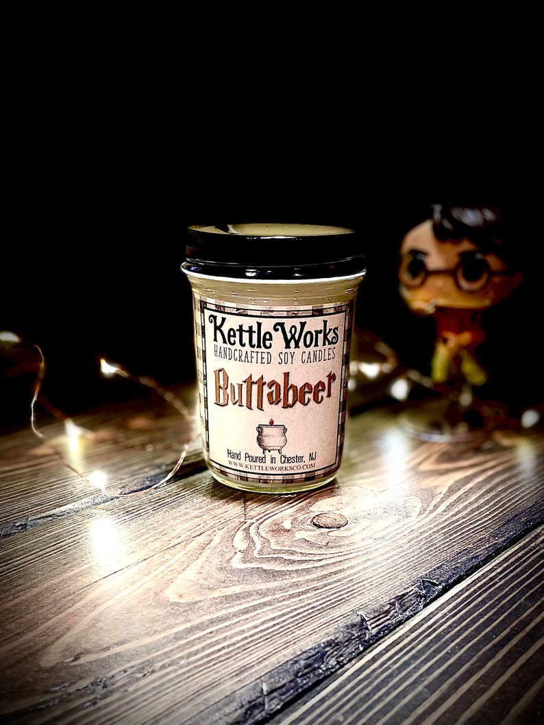May include: A glass jar candle with a black lid and a white label that says "Kettle Works Handcrafted Soy Candles Buttabeer" and "Hand Poured in Chester, NJ www.kettleworksco.com".