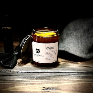 May include: A glass jar candle with a black lid, labeled "dapper." The label lists ingredients like citrus, sea salt, and vanilla. The candle is part of the Crescent Kettle Collection and is hand-poured in Chester, NJ. A black bow tie and a gray hat are in the background.