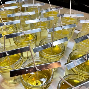 May include: A close-up of multiple glass jars filled with a clear, yellow liquid. Each jar has a metal strip across the top and a white wick extending from the liquid. The jars are arranged in rows on a white surface.