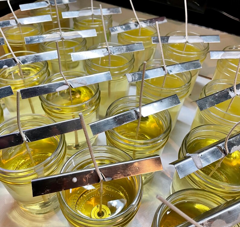May include: A close-up of multiple glass jars filled with a clear, yellow liquid. Each jar has a metal strip across the top and a white wick extending from the liquid. The jars are arranged in rows on a white surface.