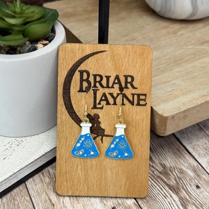 May include: A pair of blue and white enamel earrings shaped like science beakers, adorned with scientific symbols. The earrings hang from gold-colored hooks. The earrings are displayed on a wooden plaque with the text "Briar Jayne".
