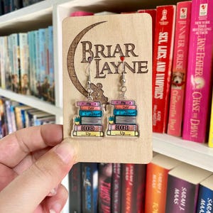 May include: A pair of book-themed earrings with a stack of books on each earring. The earrings are displayed on a wooden stand with the words "Briar Layne" engraved on it.