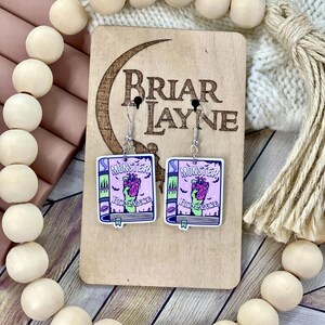 May include: A pair of earrings featuring a pink book with a green monster heart and the words "Monster Romance" on the cover. The earrings are on a wooden display stand with the words "Briar Layne" on it.