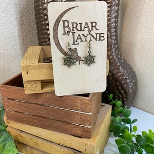 May include: A pair of bronze-colored earrings with a ship's wheel design. The earrings are displayed on a wooden card with the text "Briar Layne" engraved on it.