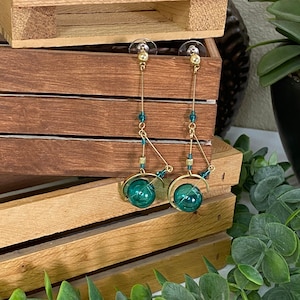 May include: A pair of long, dangling earrings with gold-toned metal and teal blue glass beads. The earrings feature a round, teal blue glass orb at the bottom.