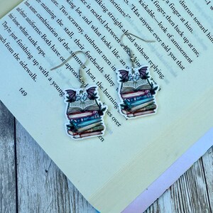 May include: A pair of dangle earrings featuring a stack of books with a bat and foliage design. The earrings have silver-colored hooks and are set against a backdrop of open book pages.