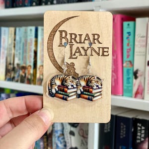 May include: A pair of cat-themed earrings with a crescent moon and the text "Briar Layne". The earrings feature a cat sleeping on a stack of books.