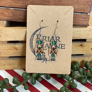 May include: A pair of silver dangle earrings featuring a green, red, and gold nutcracker design. The earrings are on a brown card with the text "PRIAR LANE" printed on it.