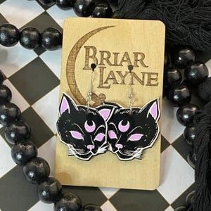 May include: A pair of black cat earrings with pink crescent moons on a wooden display card with the text "Briar Layne".
