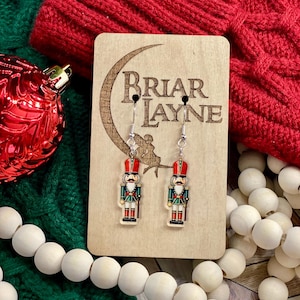 May include: A pair of nutcracker earrings on a wooden display card with the text "Briar Layne". The earrings are silver with a red, green, and white nutcracker design.
