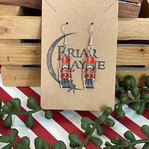 May include: A pair of silver dangle earrings featuring a red and silver nutcracker design. The earrings are on a brown card with the text "Priar Layne".