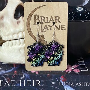 May include: A pair of black cat earrings with a pentagram, the words "Hail Satan" and "666" in rainbow colors, and the word "Meow" in black. The earrings are on a wooden display card with the words "Briar Layne" printed on it.