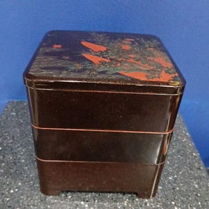 May include: A dark brown, three-tiered Japanese bento box with a square shape. The lid features a faded design with red and green accents. The box is made of a glossy material and sits on a speckled surface.