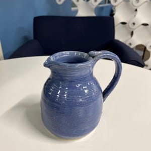 May include: A handmade, blue ceramic pitcher with a curved handle and a pouring spout. The pitcher has a textured surface and a rounded body. The pitcher is sitting on a white surface.