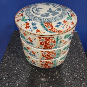 May include: A stacked, round ceramic container with a lid. The container features a white base with floral and geometric patterns in red, green, and blue. The lid has a blue and white design with birds and floral motifs. The container is on a dark surface.