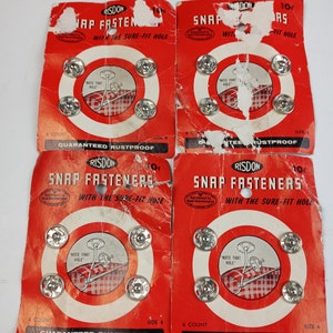 Puede incluir: Cuatro tarjetas de botones a presión plateados, cada tarjeta tiene un círculo rojo con un blanco en el centro. Las tarjetas están etiquetadas "Risdon Snap Fasteners with the Sure-Fit Hole" y "Guaranteed Rustproof".