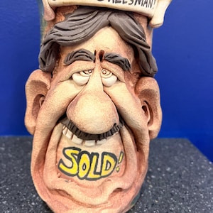 Op de afbeelding: Een keramische mok in de vorm van een cartoon gezicht met een brede glimlach en de woorden "SUPER SALESMAN!" boven het hoofd en "SOLD!" over de mond. De mok is geschilderd in tinten bruin, geel en blauw.