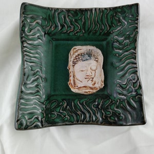 Puede incluir: Un plato cuadrado de cerámica con un esmalte verde y un diseño en relieve. El plato tiene una cabeza de Buda de cerámica blanca en el centro.