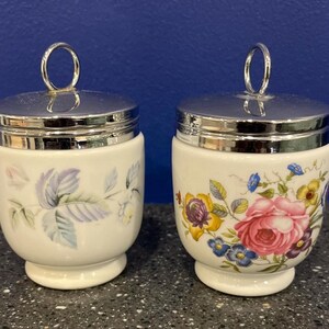 May include: A pair of vintage egg cups with floral patterns. Each has a white base and a silver-coloured lid with a ring handle. One cup displays a pink rose and other flowers, the other a lavender and green leaf design.