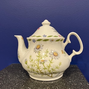 May include: White teapot with a floral design featuring daisies and other wildflowers. The teapot has a lid and a handle. The teapot is on a dark surface.