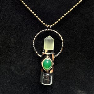 May include: A pendant necklace featuring a clear, hexagonal crystal point set within a twisted metal circle. The cylindrical base is accented with a smooth, oval-shaped green stone. The entire piece hangs from a thin, metallic ball-chain necklace.