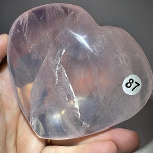 May include: A heart-shaped rose quartz stone with a smooth, polished surface and visible internal crystalline fractures. The translucent light pink mineral features a small white circular sticker with the black number 87 on its front.