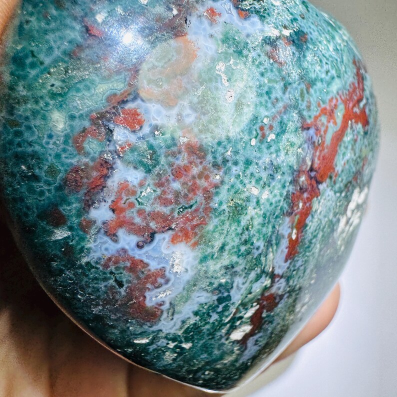 May include: A heart-shaped stone with a polished surface. The stone features a mix of colors, including deep green, red, and white. The pattern is swirled and mottled, creating a unique and natural appearance. The stone is held in a hand.
