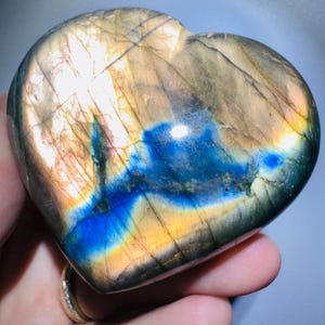 May include: A heart-shaped labradorite stone with a polished surface. The stone displays a mix of colors, including blues, yellows, and browns. The heart is held in a hand, showcasing its size and shape. The stone's unique patterns and colors are highlighted.