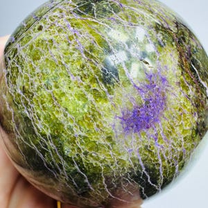 May include: A polished sphere with a mottled green and purple pattern. The surface has a network of fine, light-colored lines. The sphere's colors range from olive green to deep purple, creating a unique and natural appearance. The sphere is a decorative item.