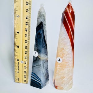 May include: Two polished stone obelisks next to a wooden ruler. The left obelisk, labeled 3, is gray, black, and white with banded patterns. The right obelisk, labeled 4, is orange, red, and white with translucent layers. The ruler shows a height of 6 inches.