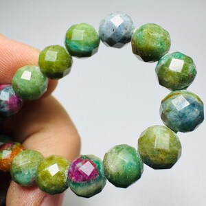 May include: A beaded bracelet featuring round, faceted gemstone beads in shades of green, blue, and occasional deep pink. The beads are arranged in a circular pattern, showcasing a polished, reflective surface with natural color variations.