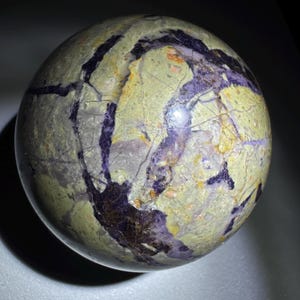 May include: A polished spherical stone featuring a mottled light green and olive base color with prominent, deep purple veins running throughout the surface. The sphere has a smooth, reflective texture with visible internal crystalline inclusions.