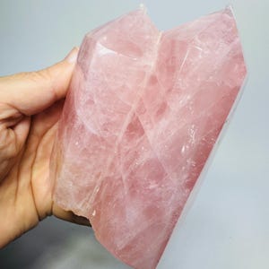 May include: A large, translucent rose quartz crystal specimen featuring two distinct, pointed terminations joined at the base. The mineral displays a soft, pale pink hue with visible internal fractures, natural inclusions, and a polished, multifaceted surface texture.