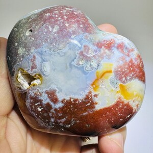 May include: A polished, heart-shaped stone with a mix of colors. The stone features areas of deep red, white, blue, and yellow. The surface has a glossy finish, and a small cavity is visible. The stone is held in a hand.