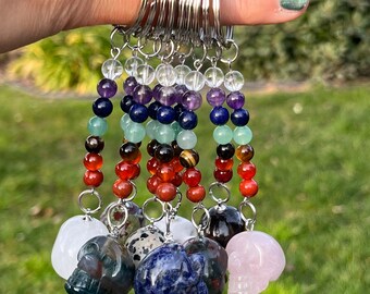 May include: A collection of keychains featuring skull pendants made from various gemstones. Each keychain has a beaded strand with chakra colors, and a silver keyring. The gemstones include clear quartz, amethyst, and rose quartz.