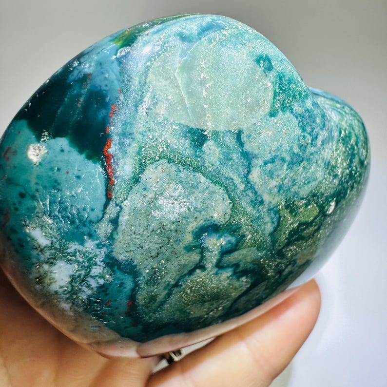 May include: A polished, heart-shaped stone featuring a marbled pattern of deep forest green, teal, and light mint green hues. The surface displays translucent areas, subtle crystalline inclusions, and a thin, distinct streak of bright red.